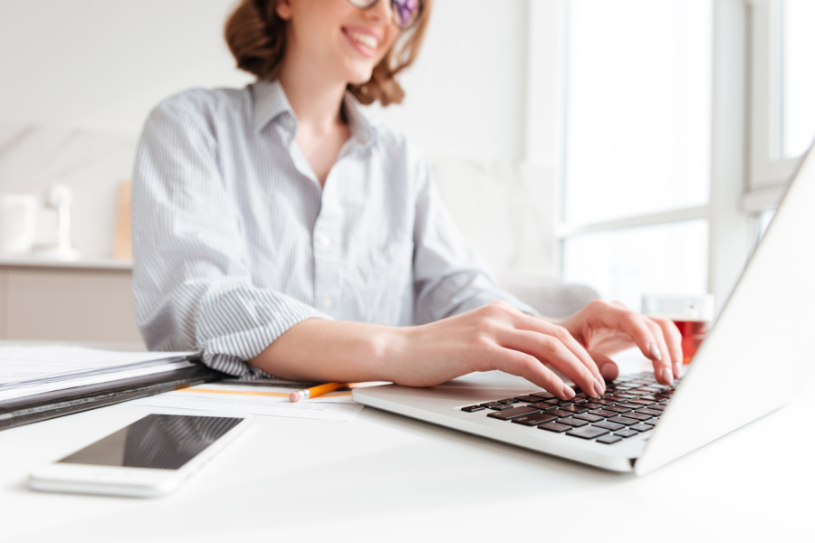 	Kobieta w okularach, uśmiechnięta, siedząca przy biurku i pisząca na laptopie. Na biurku leży smartfon, ołówek i dokumenty. W tle rozmyte elementy wnętrza.