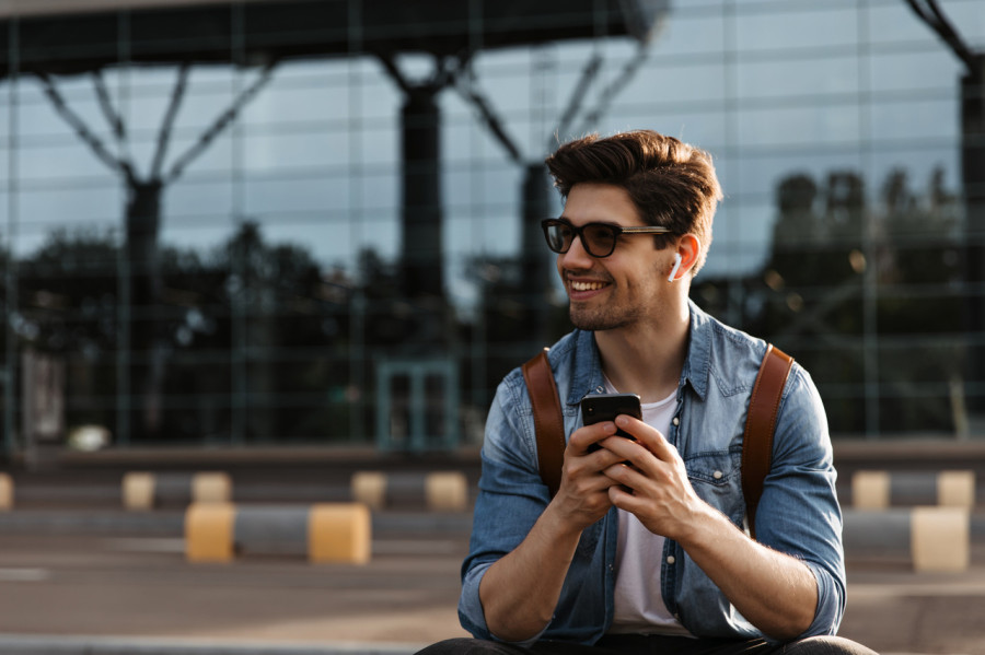 	Mężczyzna w okularach przeciwsłonecznych, uśmiechnięty, siedzący na zewnątrz przed nowoczesnym budynkiem, trzymając smartfon. Ma na sobie dżinsową koszulę i nosi plecak.